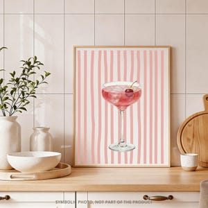 May include: A framed print featuring a watercolor illustration of a pink cocktail in a coupe glass, garnished with a cherry and lemon peel. The background has vertical pink stripes on a white backdrop.
