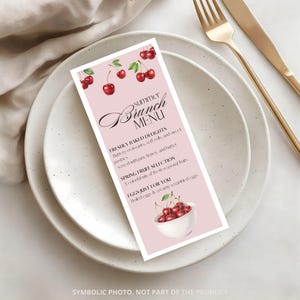May include: A pink brunch menu card with cherry illustrations, placed on a white plate with gold cutlery. The menu features text for brunch delights, fruit selection, and eggs. The card has a decorative script font for "Brunch Menu".