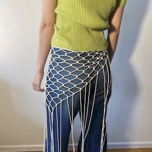 May include: A cream-colored macrame skirt with a diamond net pattern and long fringe. The skirt is worn over blue jeans. The person is wearing a green ribbed top.