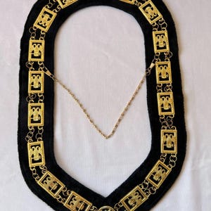Puede incluir: Un collar ceremonial dorado con respaldo de terciopelo negro. El collar presenta eslabones rectangulares con diseños intrincados y una cadena central. El diseño incluye motivos de estrellas y águilas, lo que sugiere un tema fraternal o ceremonial.