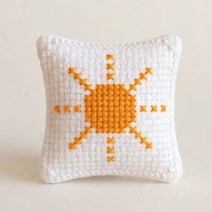 May include: A small, white, square pillow with a cross-stitch design of a yellow sunburst. The sun has a circular center and radiating lines. The pillow is a decorative item.