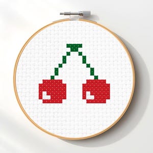 Può includere: Un telaio da ricamo a punto croce con un disegno pixelato di due ciliegie rosse con steli verdi. Le ciliegie sono realizzate con filo rosso su uno sfondo di tessuto bianco, inserite in un telaio di legno.