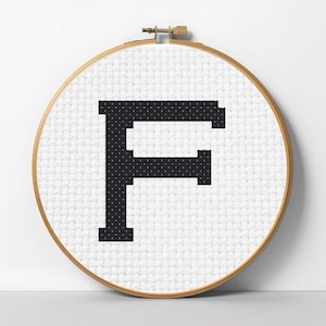 Könnte beinhalten: Ein Kreuzstich-Stickrahmen mit dem schwarzen Buchstaben "F" auf weißem Stoff. Der Buchstabe ist mittig in dem runden Rahmen platziert, der einen Holzrahmen hat. Das Design ist schlicht und grafisch.