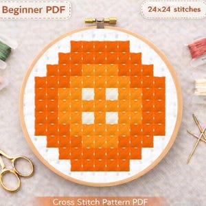 Puede incluir: Un patrón de punto de cruz en un aro de madera presenta un diseño naranja pixelado con cuadrados blancos. Los textos "Beginner PDF" y "24x24 stitches" son visibles. Las palabras "Cross Stitch Pattern PDF" están en la parte inferior.