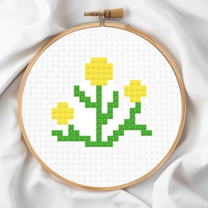 Pode incluir: Um bastidor de bordado em ponto cruz com um desenho de flores amarelas com caules e folhas verdes. As flores são em estilo pixelizado. O bordado é colocado sobre um fundo de tecido branco e o bastidor é de madeira.