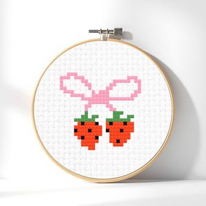 Op de afbeelding: Een borduurring met kruissteek met een ontwerp van twee rode aardbeien met groene toppen, verbonden door een roze strik. De borduursel staat tegen een witte achtergrond in een houten ring.