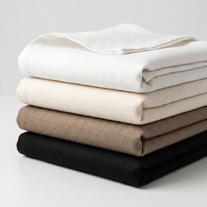 May include: A stack of four folded square fabric napkins in a gradient of colors. The top napkin is bright white, followed by a cream-colored napkin, a medium brown napkin, and a solid black napkin at the bottom, all featuring a visible woven texture.