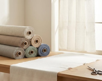 May include: A stack of six fabric rolls in neutral and muted tones including beige, gray, green, and blue, alongside a white fabric runner draped on a wooden table. A white curtain hangs on a rod in the background, and metal scissors rest nearby.