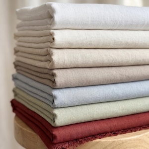 Può includere: Una pila di tessuti piegati in vari colori, tra cui bianco, crema, beige, azzurro, verde salvia e rosso scuro. I tessuti presentano una trama visibile e il pezzo inferiore mostra una finitura con bordo sfrangiato.