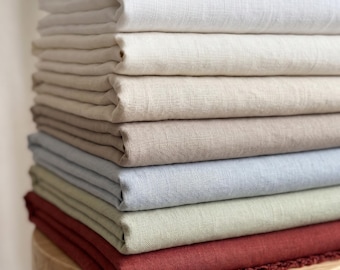 Puede incluir: Una pila de telas dobladas en varios colores, incluyendo blanco, crema, beige, azul claro, verde salvia y rojo oscuro. Los textiles presentan una textura tejida visible y la pieza inferior muestra un acabado de borde deshilachado.