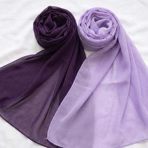 May include: Two rectangular scarves made of a lightweight, semi-sheer fabric. One scarf is a deep, dark plum purple, while the other is a lighter, soft lavender shade. Both scarves are arranged in loose, circular folds at the top with draped ends.