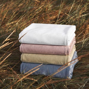 May include: A stack of four folded rectangular blankets in solid colors. The top blanket is white, followed by a dusty rose pink, a muted tan, and a light blue blanket at the bottom. Each blanket has a smooth, soft-textured surface finish.