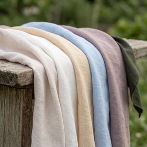 May include: A collection of folded fabric napkins in a variety of colors including white, cream, light blue, dusty rose, and dark olive green. The textiles feature a visible woven texture and are layered neatly to display each distinct color shade.
