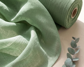 May include: A roll of sage green fabric with a visible woven texture, partially unrolled to reveal soft, draped folds. The textile features a semi-sheer quality and a consistent light green hue throughout the material, accompanied by a eucalyptus sprig.