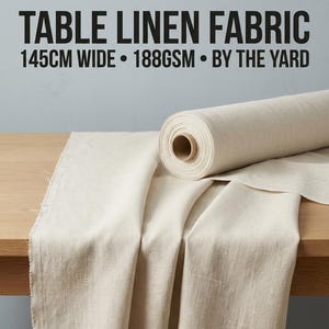 May include: A roll of off-white table linen fabric with a visible woven texture. The text on the image reads: TABLE LINEN FABRIC, 145CM WIDE, 188GSM, BY THE YARD. The fabric is draped over a wooden surface, showing its natural, light beige color.