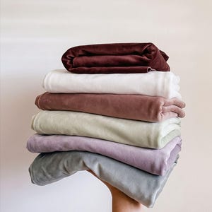 May include: A stack of six folded velvet blankets in various colors, including deep burgundy, crisp white, dusty rose, soft sage green, muted lavender, and slate gray. Each blanket has a smooth, soft texture and is neatly layered in a vertical pile.