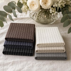 May include: A collection of six folded fabric swatches arranged in two stacks. The left stack features dark brown, navy blue, and black fabrics, while the right stack includes cream, off-white, and gray fabrics, all with a white grid pattern.
