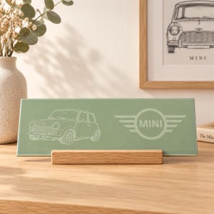 May include: A rectangular sage green ceramic tile featuring a white line drawing of a classic Mini car on the left and the Mini logo on the right. The tile is supported by a light-coloured wooden stand, displaying a clean and simple design.