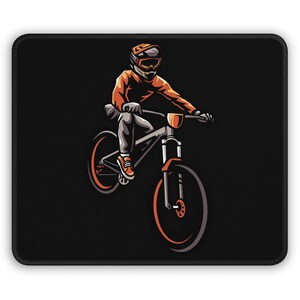 Könnte beinhalten: Schwarzes Mauspad mit einer stilisierten Illustration eines Mountainbikers in orangefarbener und grauer Ausrüstung. Der Biker fährt ein Mountainbike mit orangefarbenen Akzenten. Der Hintergrund ist einfarbig schwarz.