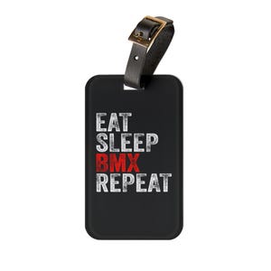 May include: Black luggage tag with the text "EAT SLEEP BMX REPEAT" in white and red. The tag has a black strap with a gold-coloured buckle.