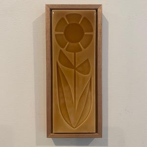 May include: A rectangular, ceramic tile with a sunflower design in shades of brown, set within a wooden frame. The sunflower is depicted with a central circle and petal shapes. The tile is mounted on a white wall.