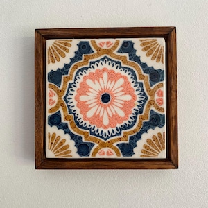May include: Square decorative tile with a floral design in a wooden frame. The tile features a symmetrical pattern with a pink, navy blue, and gold colour scheme. The frame is a dark brown colour.