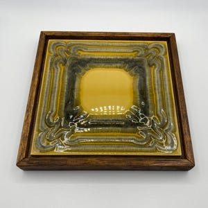 May include: A square, decorative tile with a yellow center and a patterned border in shades of yellow, gray, and brown. The tile is set in a dark brown wooden frame, creating a vintage aesthetic.