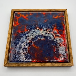 Puede incluir: Azulejo cerámico cuadrado con un diseño abstracto azul oscuro y rojo, enmarcado en un marco de madera natural. La superficie del azulejo tiene un aspecto texturizado con una mezcla de colores. El marco de madera añade un toque rústico.