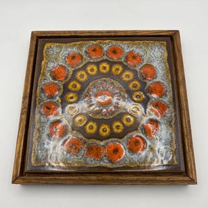 May include: Square ceramic tile with a brown, orange, and yellow floral design, set in a wooden frame. The tile features a central circular pattern surrounded by orange flower-like shapes.