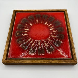 May include: A square, red ceramic tile with a brown and white floral design, set in a wooden frame. The tile has a glossy finish and a raised, textured pattern. The wooden frame is a warm, medium brown colour.