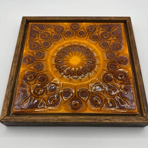 May include: Square ceramic tile with a brown wooden frame. The tile features a repeating pattern of brown and orange floral designs, with a central sunburst pattern in the center. The tile has a glossy finish.