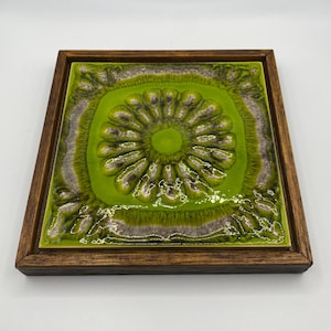 May include: A square, framed ceramic tile with a vibrant green floral design. The center features a raised, circular pattern, surrounded by a textured, light green and gray border. The frame is made of dark brown wood.