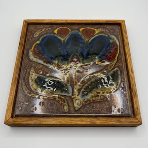Puede incluir: Azulejo de cerámica cuadrado con un diseño floral en un marco de madera. La flor tiene pétalos azules, rojos y amarillos con hojas verdes. El azulejo tiene un fondo marrón y un acabado brillante.