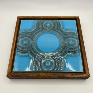 Puede incluir: Una baldosa de cerámica cuadrada con un fondo azul claro y un diseño circular en capas en tonos azules y marrones. La baldosa está enmarcada en un marco de madera marrón oscuro, creando una pieza de arte decorativa.