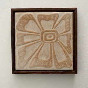 May include: A square, framed tile featuring a stylized flower design. The flower has a central circle with radiating petals in shades of tan and brown. The tile is set within a dark brown wooden frame, creating a decorative wall art piece.