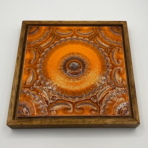 May include: Square orange ceramic tile with a brown and white circular design, set in a wooden frame. The tile features a repeating paisley-like pattern around the central circle. The frame is made of light brown wood.
