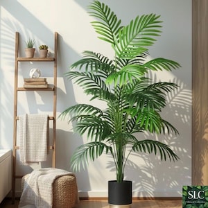 Puede incluir: Una gran palmera artificial con exuberantes hojas verdes en una maceta negra con base dorada. La planta se coloca junto a una estantería de escalera de madera con pequeñas plantas en macetas y un otomán tejido con una manta. El escenario es contra una pared blanca.