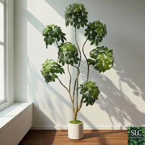 Puede incluir: Una planta artificial alta con múltiples tallos y grandes grupos de hojas verdes. La planta está en una maceta cilíndrica blanca y se encuentra en un piso de madera. El fondo es una pared blanca con luz solar.