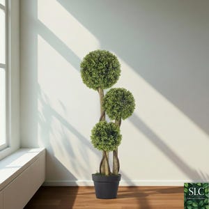 Könnte beinhalten: Ein künstlicher Topiari-Baum mit drei kugelförmigen grünen Blattgruppen. Der Baum steht in einem dunkelgrauen Topf und hat einen natürlich aussehenden Stamm. Das Bild ist vor einer weißen Wand mit Sonnenlicht durch ein Fenster aufgenommen.