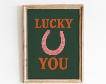 May include: A framed print with the words "LUCKY YOU" in orange letters on a dark green background. A pink horseshoe is centered between the words. The frame is a light brown color.