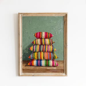May include: An oil painting in a wooden frame depicting a stack of five rectangular pillows with vertical stripes in vibrant colors including pink, orange, yellow, green, blue, and purple, set against a textured sage green background.