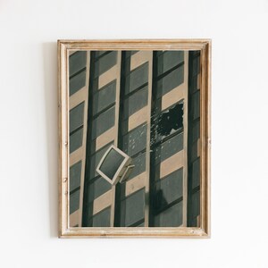 May include: A framed wall art print depicting a tall building facade with dark rectangular windows and light-coloured vertical panels. A single white computer monitor is suspended in front of the glass, appearing to float against the building exterior.