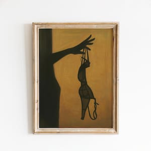 Può includere: Opera d'arte incorniciata con la silhouette di una mano che tiene un reggiseno. Il reggiseno è scuro su uno sfondo caldo e dorato. L'opera d'arte è presentata in una cornice di legno.