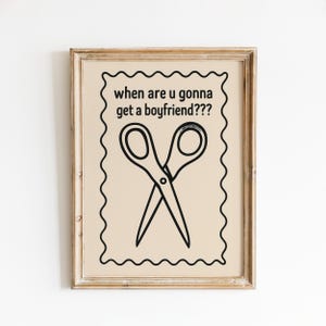 May include: A framed print with the text "when are u gonna get a boyfriend???" above a black and white illustration of crossed scissors. The print has a beige background and a decorative border, set in a light brown wooden frame.
