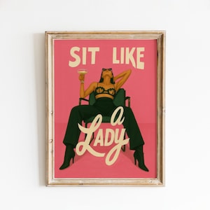 May include: Framed art print with a pink background and the words "SIT LIKE A LADY" in white. The image features a woman in a chair, wearing a black bra and trousers, holding a cocktail. The art print is in a gold frame.