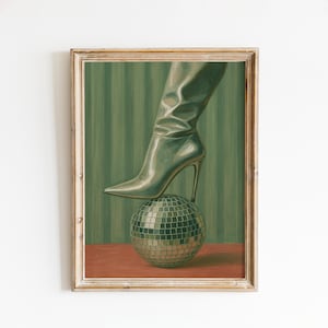 May include: Framed artwork featuring a silver high-heeled boot resting on a disco ball. The background has vertical green stripes, and the base is a reddish-brown color. The artwork is in a gold-colored frame.