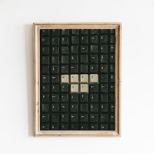 May include: A framed wall art print featuring a grid of black computer keyboard keys with directional arrows. Six light-coloured keys in the centre spell out the words WAKE UP in black capital letters, contrasting against the dark background.
