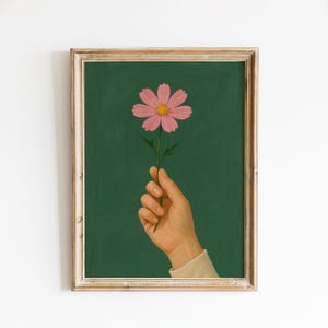 May include: A framed art print featuring a pink cosmos flower held by a hand against a dark green background. The flower has a yellow center and is on a green stem. The frame is a light wood color.