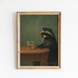 May include: A framed art print showing a raccoon smoking a cigarette and sitting at a small wooden table with a glass of amber liquid. The raccoon is depicted in a realistic style against a green backdrop. The frame is light brown.