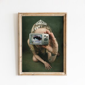 Può includere: Opera d'arte incorniciata raffigurante una persona che tiene in mano una fotocamera digitale, lo schermo mostra un primo piano di un occhio. Sulla fotocamera c'è la scritta "Self portraits". La persona indossa una tiara.